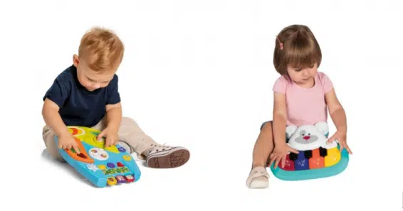 A imagem mostra à esquerda um menino de 2 anos, loiro de pele clara, sentado ao chão, segurando uma brinquedo musical. À direita temos uma menina de 2 anos, cabelos marrons e pele clara, sentada no chão, segurando seu brinquedo musical. O fundo é branco e o chão é branco também. 