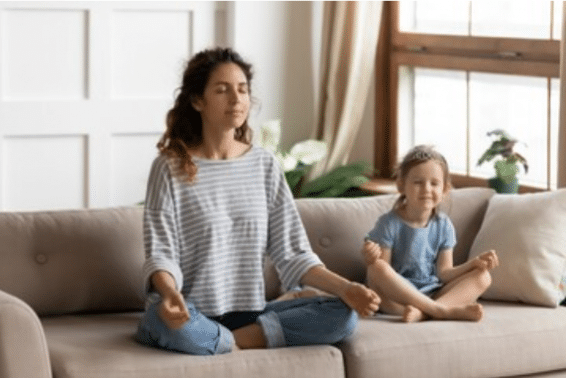 A imagem mostra uma sala, com um sofá de tecido bege, uma mãe à esquerda e uma filha à direta. Ambas estão sentadas no sofá, com as pernas cruzadas realizando uma posição de Yoga para meditação.