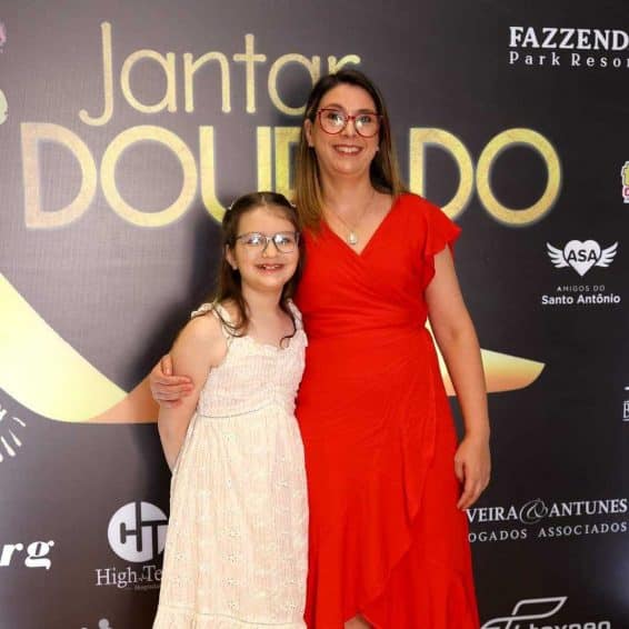 Mãe e filha, lado a lado, sorrindo para a foto. Atrás dela pode ser visto um painel de cor preta com escritas em dourado. A escrita é "Janta Dourado", uma alusão à campanha de conscientização do câncer infantil que acontece no mês de novembro e é chamado de Novembro Dourado.
