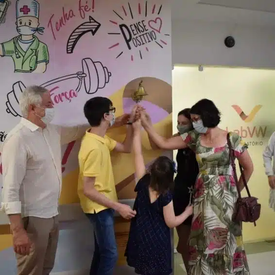 Família unida com uma criança, bateno um sino ue está preso a uma parede com mensagens de pensamento positivo, celebrando a conquista do vencimento do câncer infantil