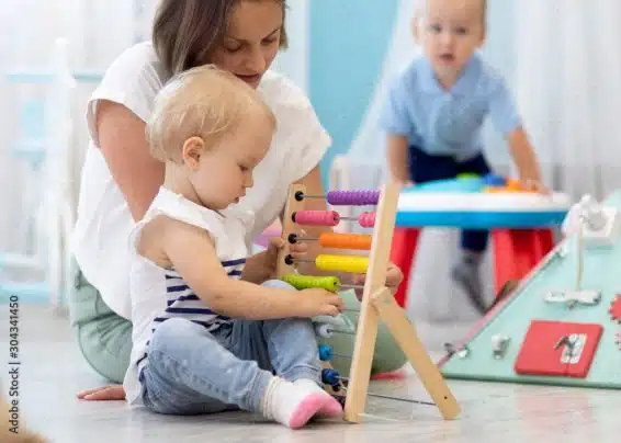 Bebê brincando com um ábaco, com o auxílio de uma professora, em uma creche, com outra criança brincando ao fundo.