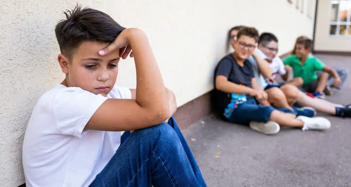 Menino triste sentado em um corredor, sofrendo bullying, com a expressão triste enquanto outras crianças riem ao fundo, olhando para o menino.