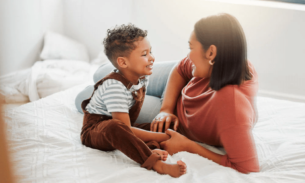Criança sorrindo para mãe, ambos em uma cama, após a cura do câncer infantil.