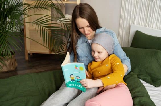 Criança com câncer sentada no sofá, lendo um livro infantil com a mãe, que está sentada ao seu lado.
