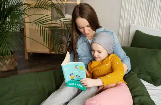 Criança com câncer sentada no sofá, lendo um livro infantil com a mãe, que está sentada ao seu lado.