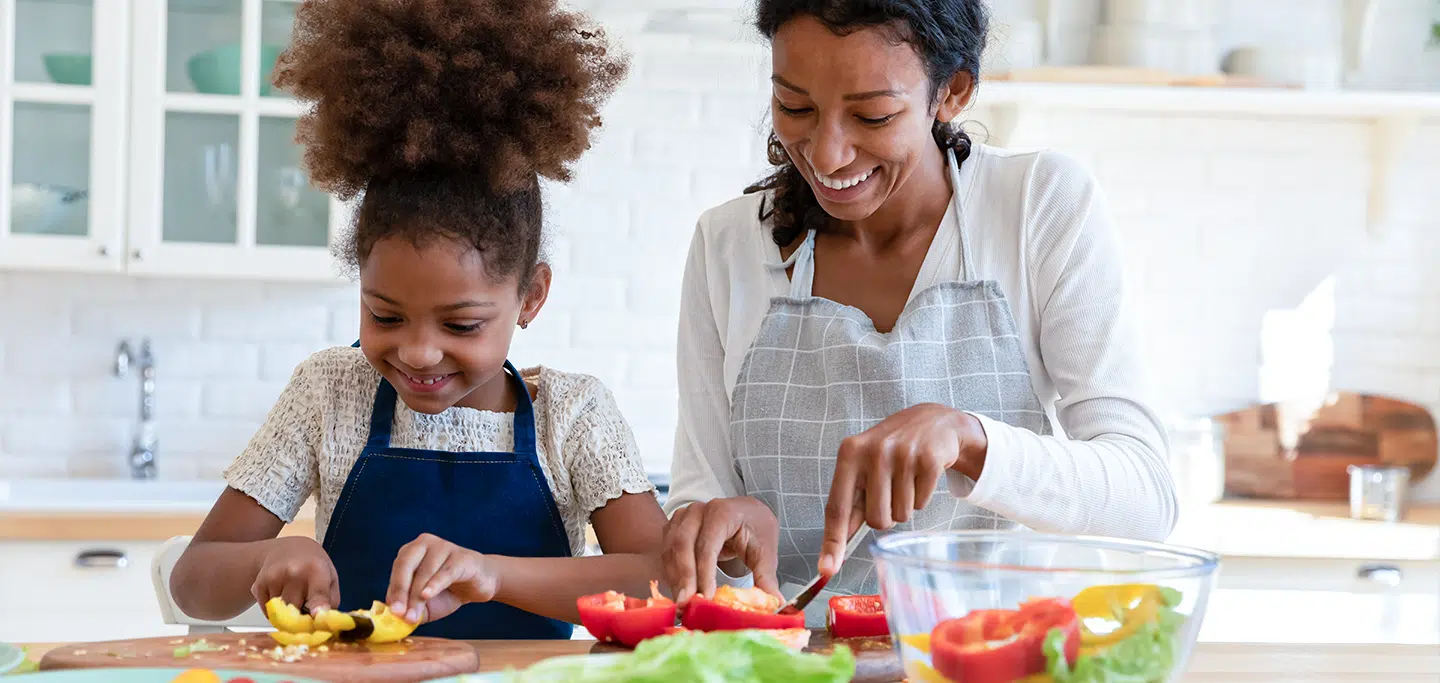 Mãe e filha criança cozinhando juntas, cortando legumes e incentivando uma alimentação saudável.