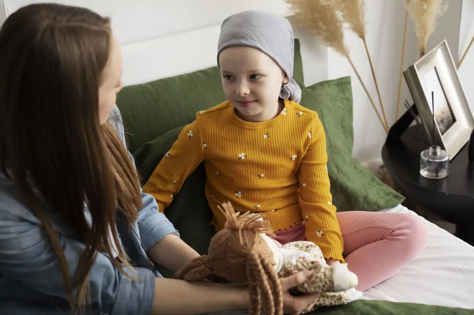 a importancia da piscologia para a criança com cancer. menina com lenço na cabeça, conversando com uma psicologa, que segura uma boneca.